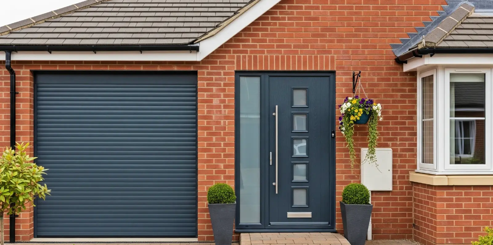 Anthracite grey roller garage door on a red brick house beside a grey front door and planters